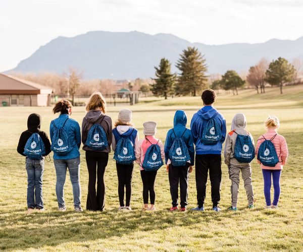 A variety of kids of all ages wearing a blue plaid TrueSport branded drawstring bag.